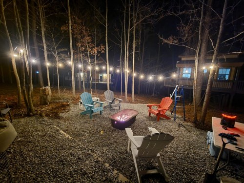 A cozy outdoor seating area with colorful chairs around a fire pit, surrounded by trees and string lights at night.