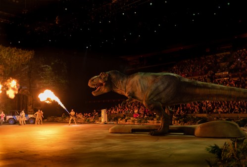 A lifelike dinosaur roars on stage, surrounded by performers and a cheering audience, with flames shooting from its mouth.