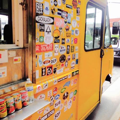 A vibrant yellow food truck covered in various colorful stickers and decals, parked on a city street.