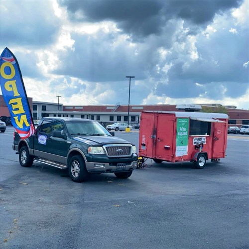 A green truck parked next to a red food trailer in a parking lot, with a colorful banner nearby.