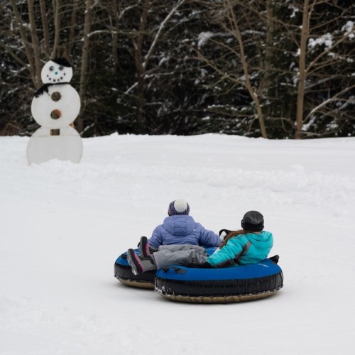 Two people in snow tubes slide down a snowy hill, with a snowman in the background.