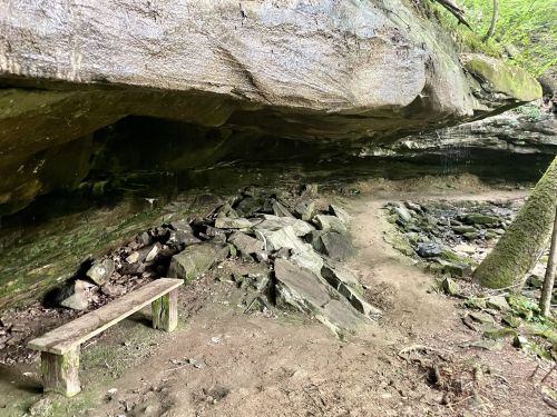 A rocky cave with a wooden bench, surrounded by greenery and natural stone formations.