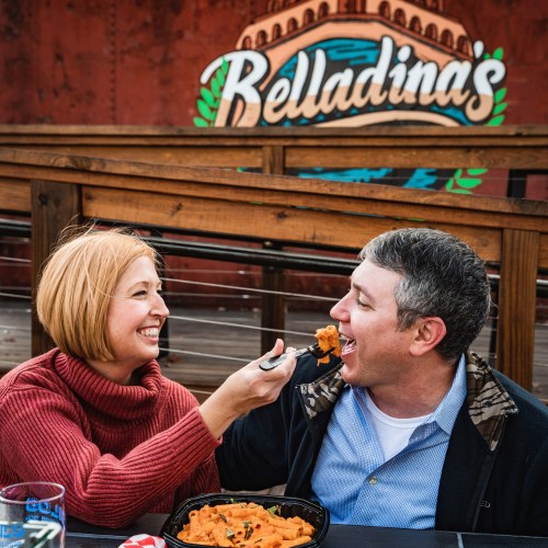 A woman playfully feeds a man pasta outdoors, with a colorful "Belladinas" sign in the background.
