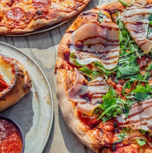 A close-up of a pizza topped with greens, prosciutto, and drizzled sauce, alongside another pizza and a stuffed crust.