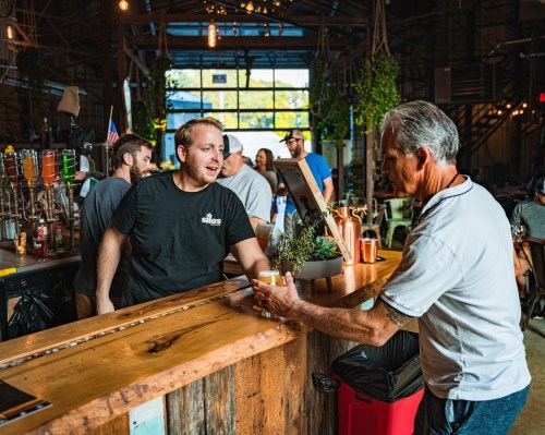 A bartender serves a drink to a customer at a lively bar with plants and a rustic wooden counter.