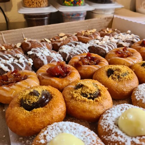 A variety of colorful donuts topped with icing, bacon, and sprinkles, arranged neatly in a box.