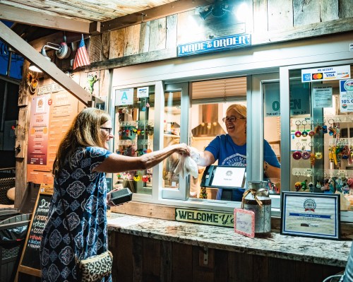 A customer receives a takeout order from a cheerful server at a food window in a rustic setting.