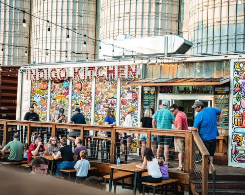 A vibrant outdoor dining area at Indigo Kitchen, with colorful murals and people enjoying food and drinks.