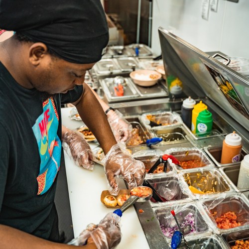 A chef prepares food in a kitchen, using gloves and various toppings from containers on the counter.