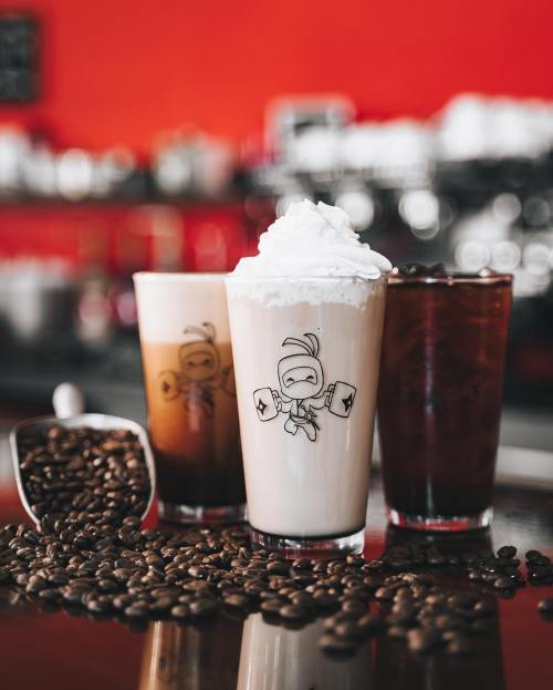 Three coffee drinks with whipped cream and a ninja logo, surrounded by coffee beans on a reflective surface.