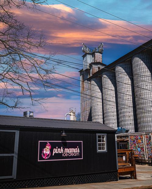 A black ice cream shop with a pink sign in front of a large grain silo under a colorful sky.