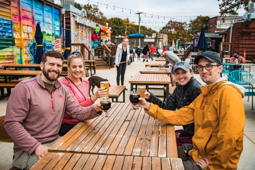 A group of four friends cheers with drinks at an outdoor venue, surrounded by colorful decor and festive atmosphere.