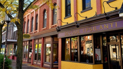 Colorful storefronts line a street in Ypsilanti, featuring a yellow building with "Decode Ypsilanti" signage.