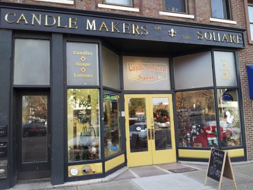 Storefront of "Candle Makers on the Square," featuring candles, soaps, and lotions, with colorful signage and decorations.