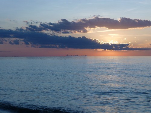 A serene sunset over calm waters, with clouds reflecting warm hues and a distant city skyline on the horizon.