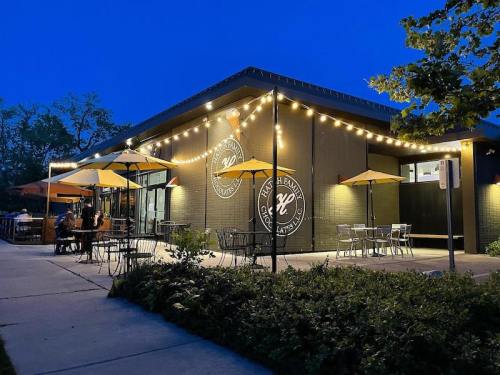 A modern restaurant exterior at dusk, featuring outdoor seating with umbrellas and warm string lights.