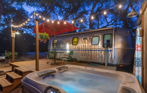 A cozy outdoor scene featuring a hot tub, an Airstream trailer, and string lights under a twilight sky.
