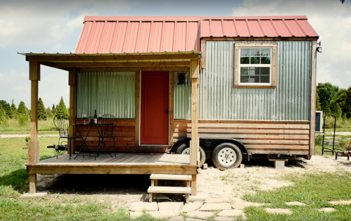 A small, rustic tiny house with a red metal roof, wooden porch, and a table set outside on a grassy area.
