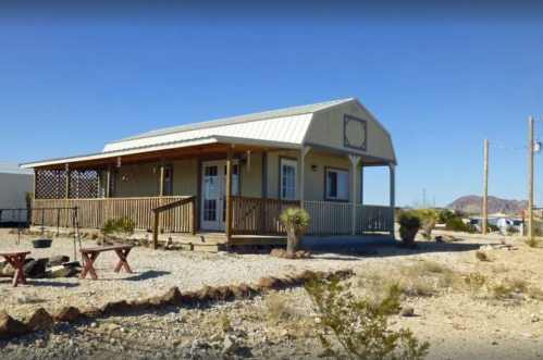 A small, modern house with a porch, surrounded by gravel and sparse vegetation in a desert landscape.