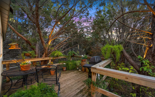 A wooden deck surrounded by trees, featuring a grill, potted plants, and a cozy seating area at dusk.
