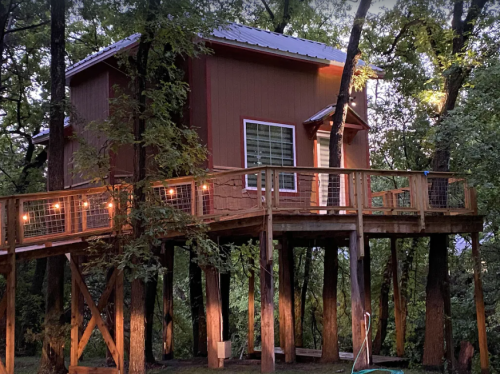 A cozy treehouse with a wooden deck, surrounded by trees and softly lit in the evening.