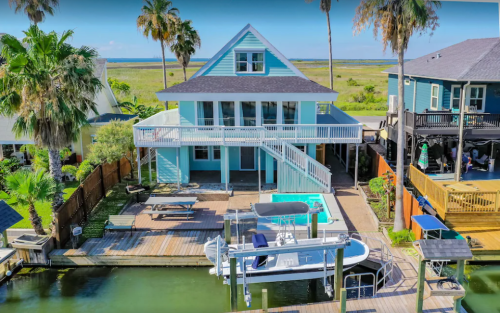 Aerial view of a blue house with a deck, surrounded by palm trees and water, featuring a boat dock and grassy landscape.
