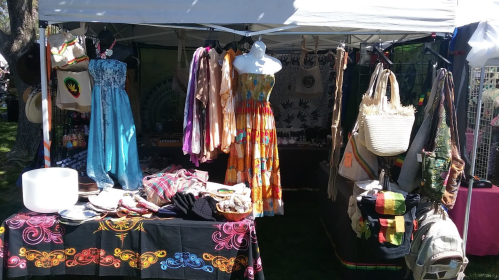 A vibrant market stall displaying colorful dresses, textiles, and handmade accessories under a white canopy.
