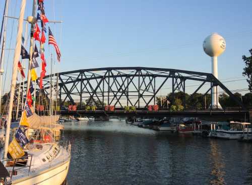 A black metal bridge spans a calm waterway, with boats docked nearby and a water tower in the background.