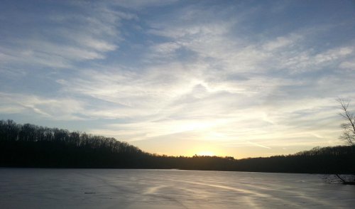 A serene winter landscape featuring a frozen lake and a sunset behind tree-lined hills under a cloudy sky.