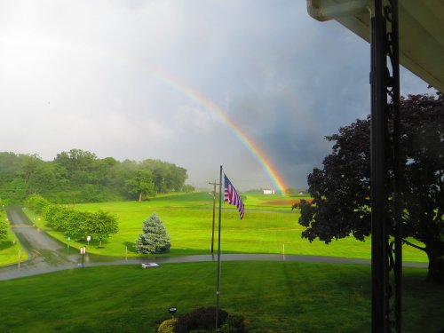 A vibrant rainbow arcs over a green landscape, with an American flag waving in the foreground after a rainstorm.