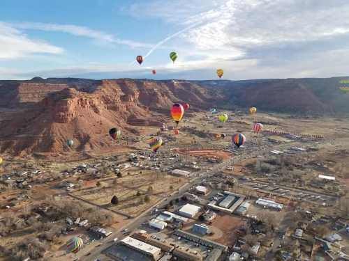 Aerial view of colorful hot air balloons floating over a valley with red rock formations and a small town below.