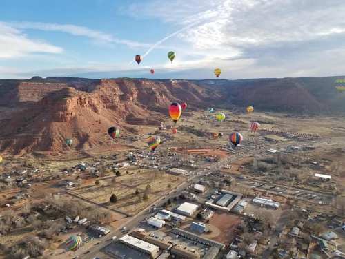 Aerial view of colorful hot air balloons floating over a valley with red rock formations and a small town below.