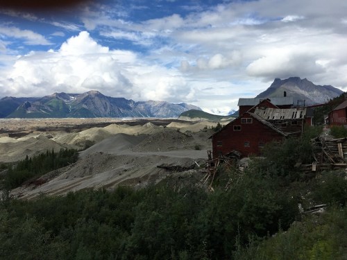 A weathered red building sits among rocky hills and mountains under a cloudy sky, surrounded by lush greenery.