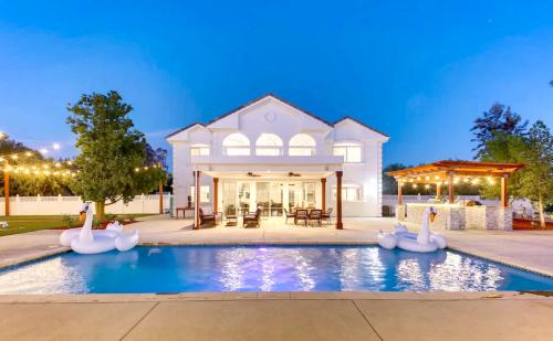 A spacious white house with a pool, swan floaties, and outdoor seating, illuminated at dusk with string lights.