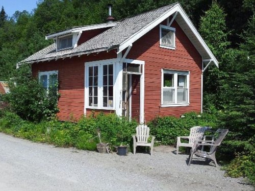A small red house with white trim, surrounded by greenery, featuring a gravel path and outdoor seating.