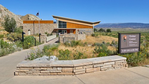 A modern visitor center with stone and wood design, surrounded by greenery and mountains, with a sign displaying hours.