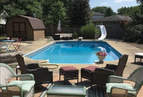 A backyard pool area with a slide, surrounded by chairs, a shed, and greenery under a clear blue sky.