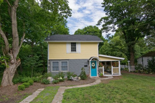 A charming two-story house with yellow and gray siding, a blue door, and a landscaped yard surrounded by trees.