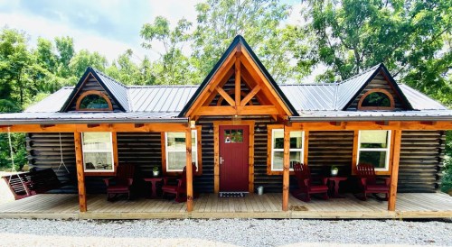 A cozy wooden cabin with a porch, featuring rocking chairs and surrounded by greenery under a blue sky.