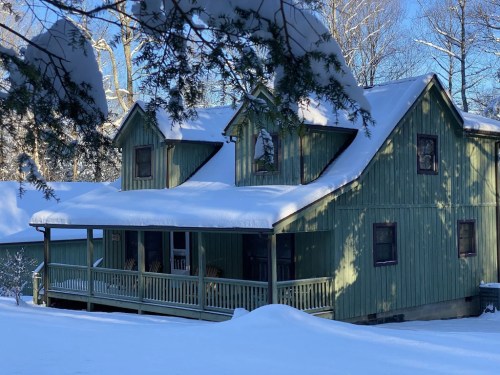 A green house with a porch, surrounded by snow-covered trees and a blanket of snow on the ground.