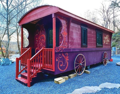 A vibrant red tiny house on wheels with decorative swirls, set in a scenic outdoor area with gravel and trees.