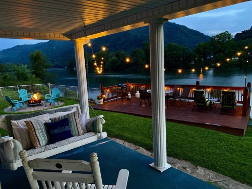 A cozy porch view at dusk, featuring a swing, string lights, and a deck overlooking a calm river and mountains.