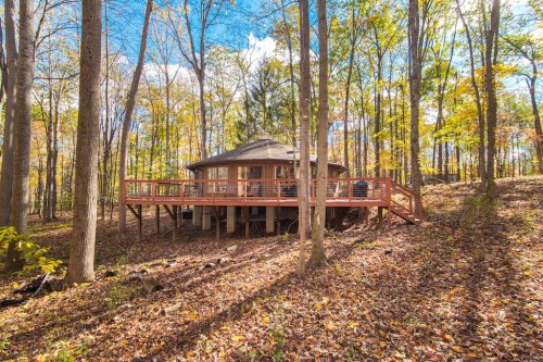 A round wooden house on stilts surrounded by trees and autumn foliage, with a deck and clear blue sky.