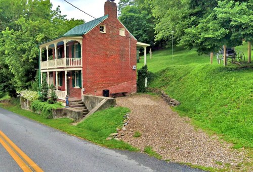 A red brick house with a green roof, surrounded by greenery, located beside a gravel driveway and a road.