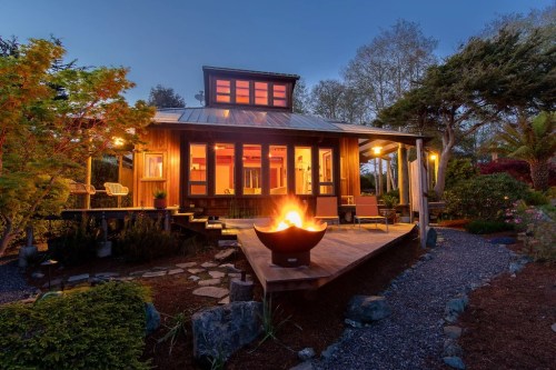 A cozy wooden house with a fire pit, surrounded by greenery, illuminated at dusk.