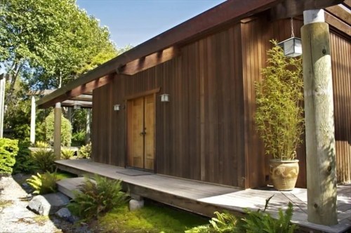 A modern wooden house with a porch, surrounded by greenery and decorative plants.
