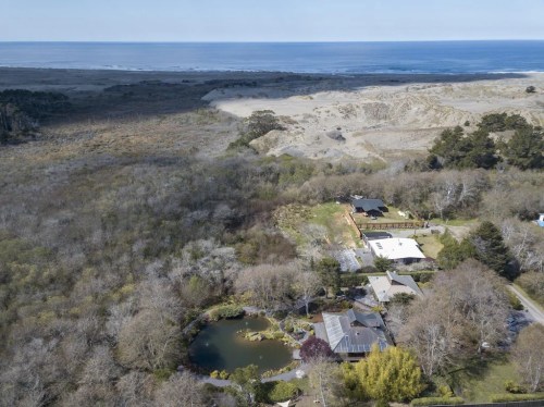 Aerial view of a coastal landscape featuring a house, pond, and surrounding trees, with the ocean in the background.