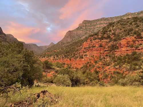 A vibrant sunset casts colorful hues over red rock formations and green vegetation in a mountainous landscape.