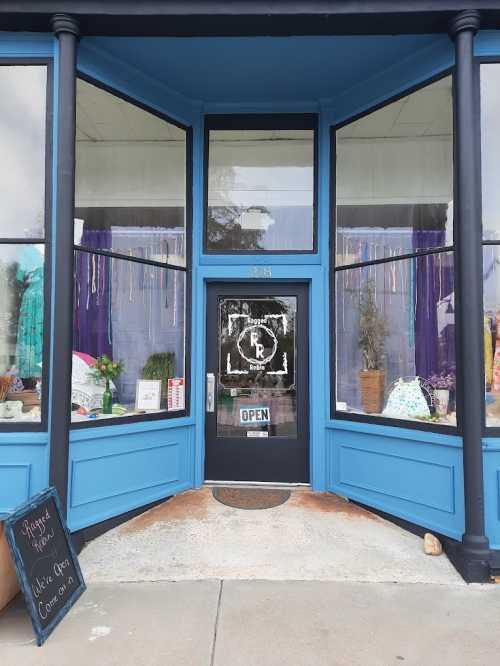 A storefront with blue walls, large windows, and an "Open" sign, featuring colorful decorations and plants inside.