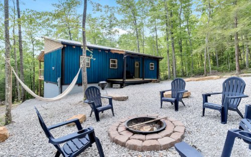A blue cabin surrounded by trees, with a fire pit and seating area in front, and a hammock hanging nearby.